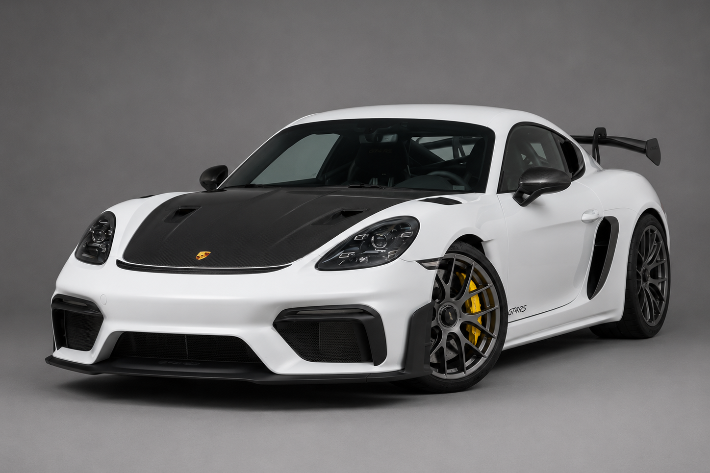 White Porsche 718 Cayman GT4 RS coupe with exposed carbon-fibre bonnet and large swan-neck rear wing, front three-quarter studio view on a plain grey backdrop showing NACA-ducted bonnet, dark forged wheels and yellow ceramic brake calipers.