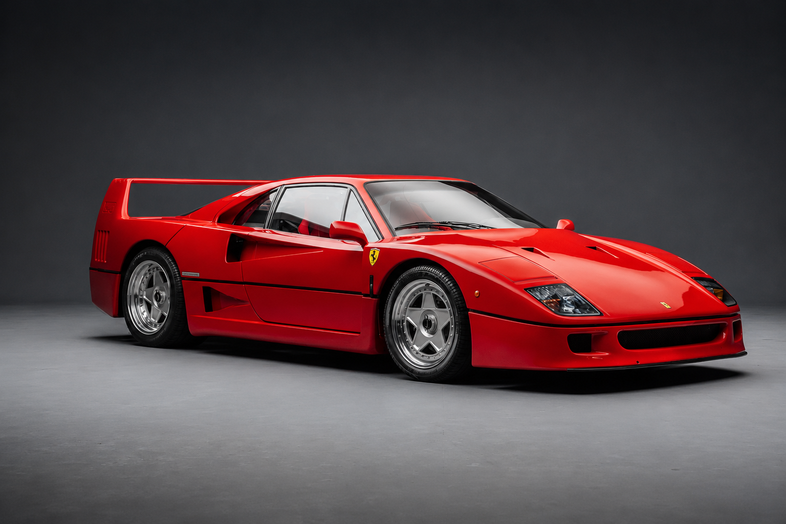 Red Ferrari F40 in a studio setting on a dark grey backdrop, front three-quarter view showing the low nose with slim covered headlamps, bonnet louvres, side NACA intakes, five-spoke alloy wheels and the fixed rear wing.