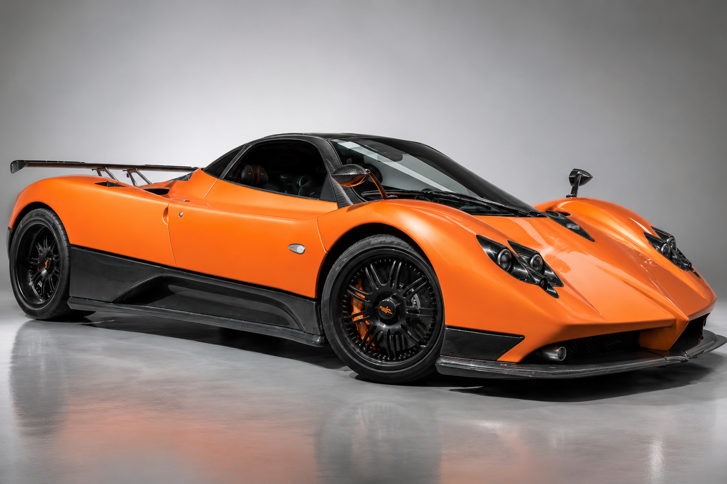 Orange Pagani Zonda in a studio, front three-quarter view showing the exposed carbon side skirts, rear wing, quad-exit exhaust and black multi-spoke wheels with orange brake calipers.