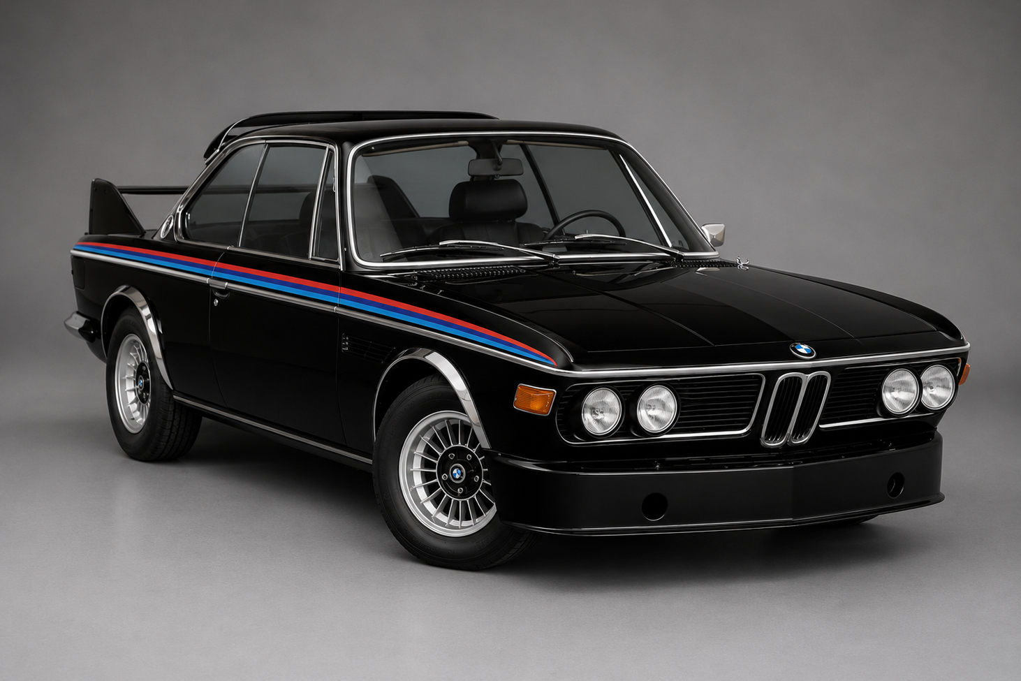 Black BMW 3.0 CSL 'Batmobile' coupe with BMW Motorsport blue, purple and red stripes along the side, rear roof spoiler and front air dam, front three-quarter studio view on a plain grey backdrop.