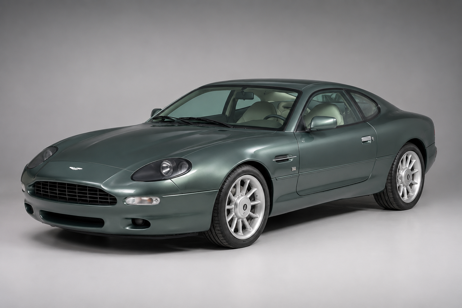 Metallic green Aston Martin DB7 coupe in a studio setting, front three-quarter view showing the smooth Ian Callum bodywork, silver multi-spoke wheels and cream leather interior.