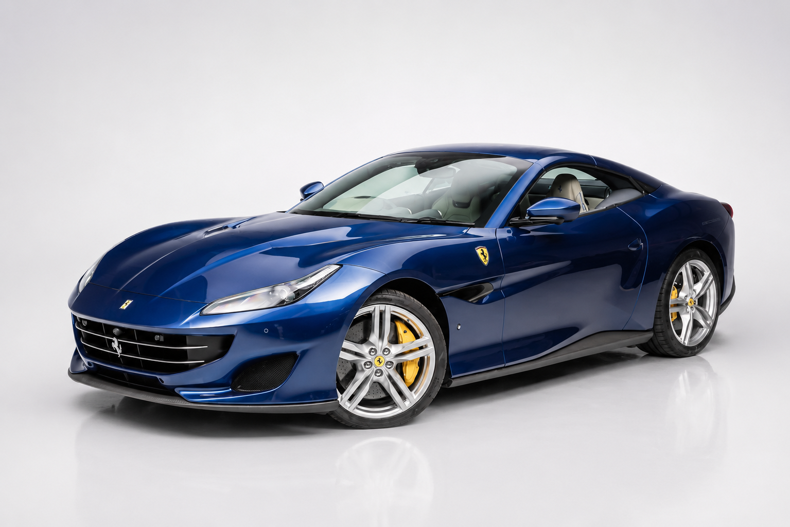 Blue Ferrari Portofino coupé photographed in a studio with the retractable hardtop raised, front three-quarter view showing the light-coloured interior, black lower grille, LED headlamps, side vents, yellow brake calipers and silver multi-spoke alloy wheels.