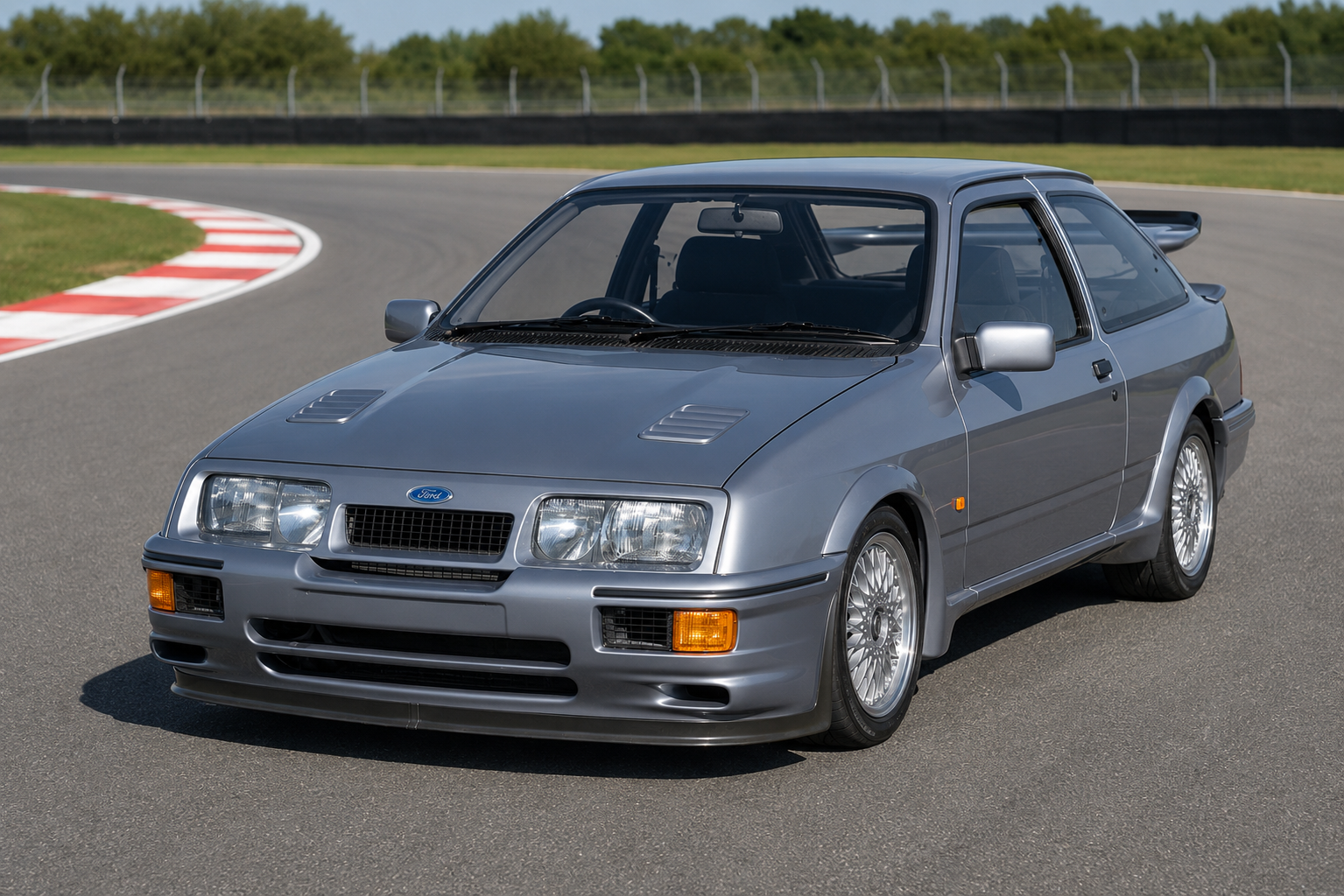 Silver Ford Sierra RS Cosworth three-door hatchback on a racetrack, front three-quarter view showing the twin bonnet vents, deep front bumper, cross-spoke alloy wheels and rear whale-tail spoiler.