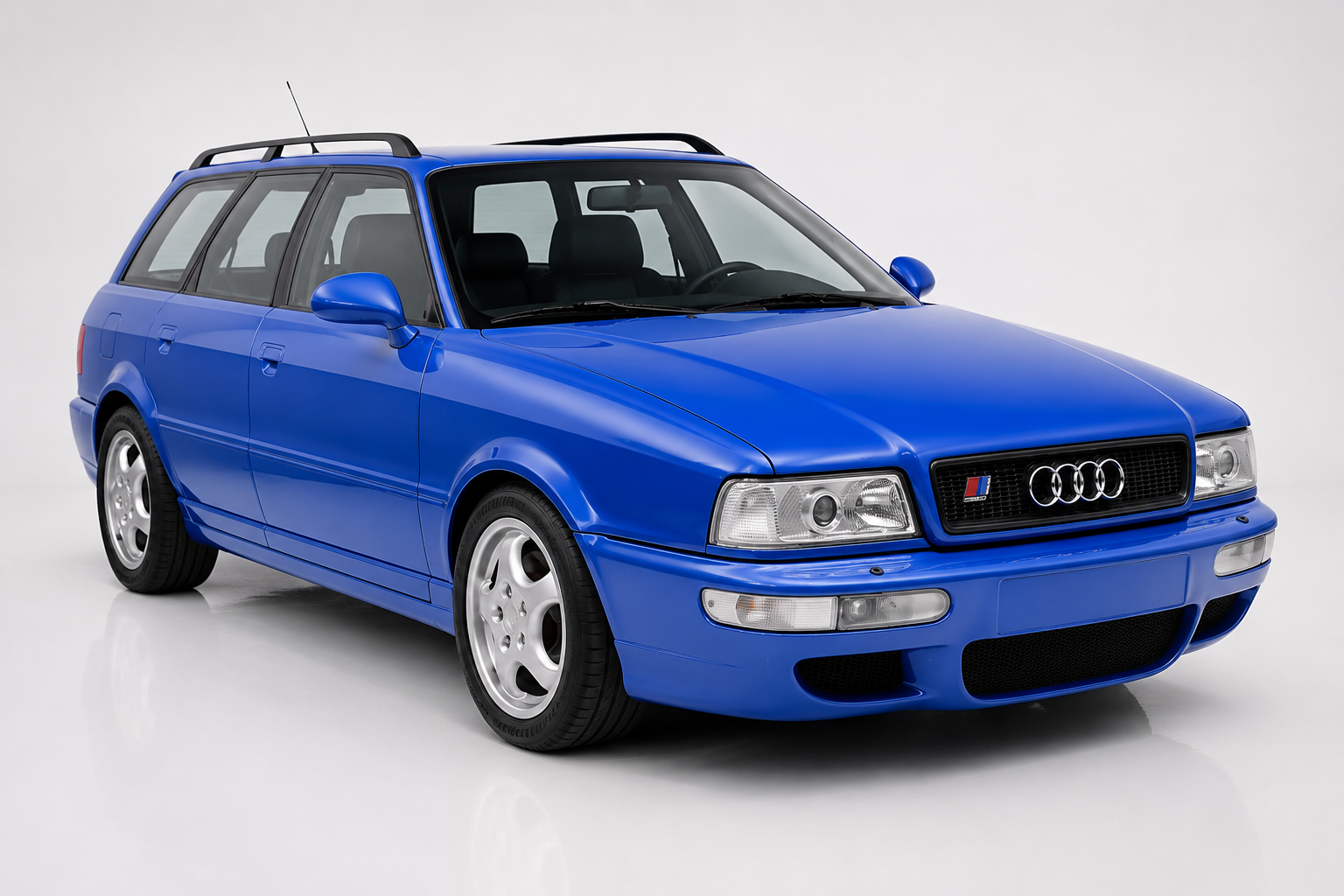 Blue Audi RS2 Avant estate in a bright studio, front three-quarter view showing the 'RS2' Porsche-badge grille, twin round headlights with amber indicators, black roof rails and silver five-spoke Porsche Cup-style alloy wheels.
