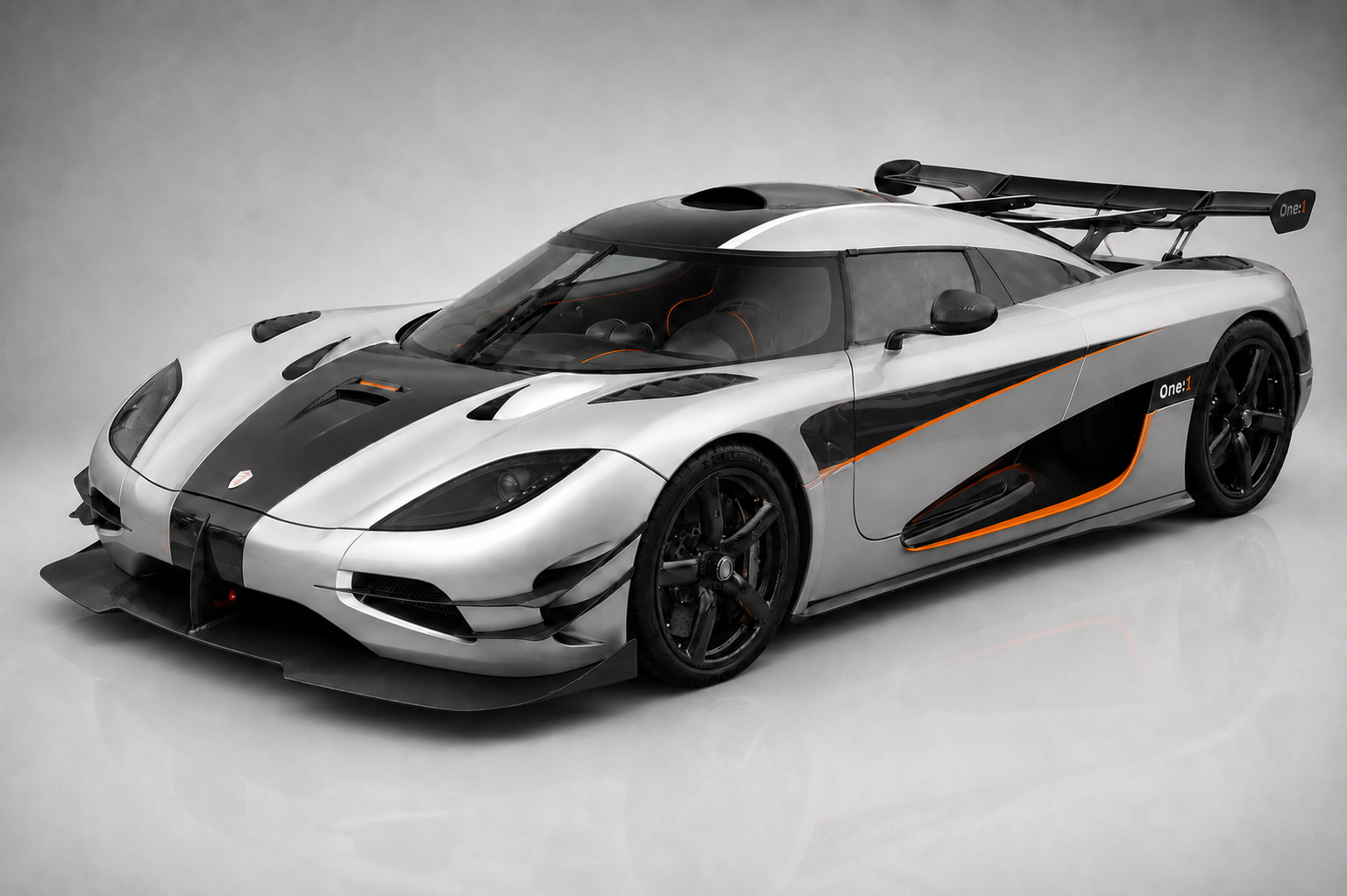 Silver and carbon-fibre Koenigsegg One:1 hypercar in a studio setting, front three-quarter view showing the twin-turbo V8 cooling intakes, aggressive front splitter and canards, distinctive 'One:1' side graphics, large rear wing and the lightweight carbon-fibre bodywork.
