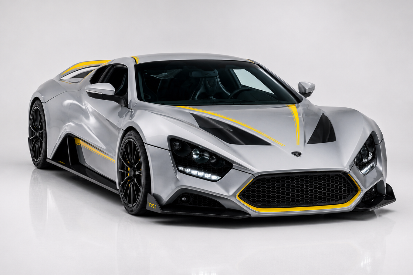 Zenvo TS1 GT in silver with contrasting yellow pinstripe accents in a bright studio setting, showing the mid-engined two-seat coupé silhouette, aggressive front splitter, exposed carbon side sills, dark multi-spoke forged wheels and the sharp front-quarter treatment of Zenvo's second-generation hand-built Danish hypercar.