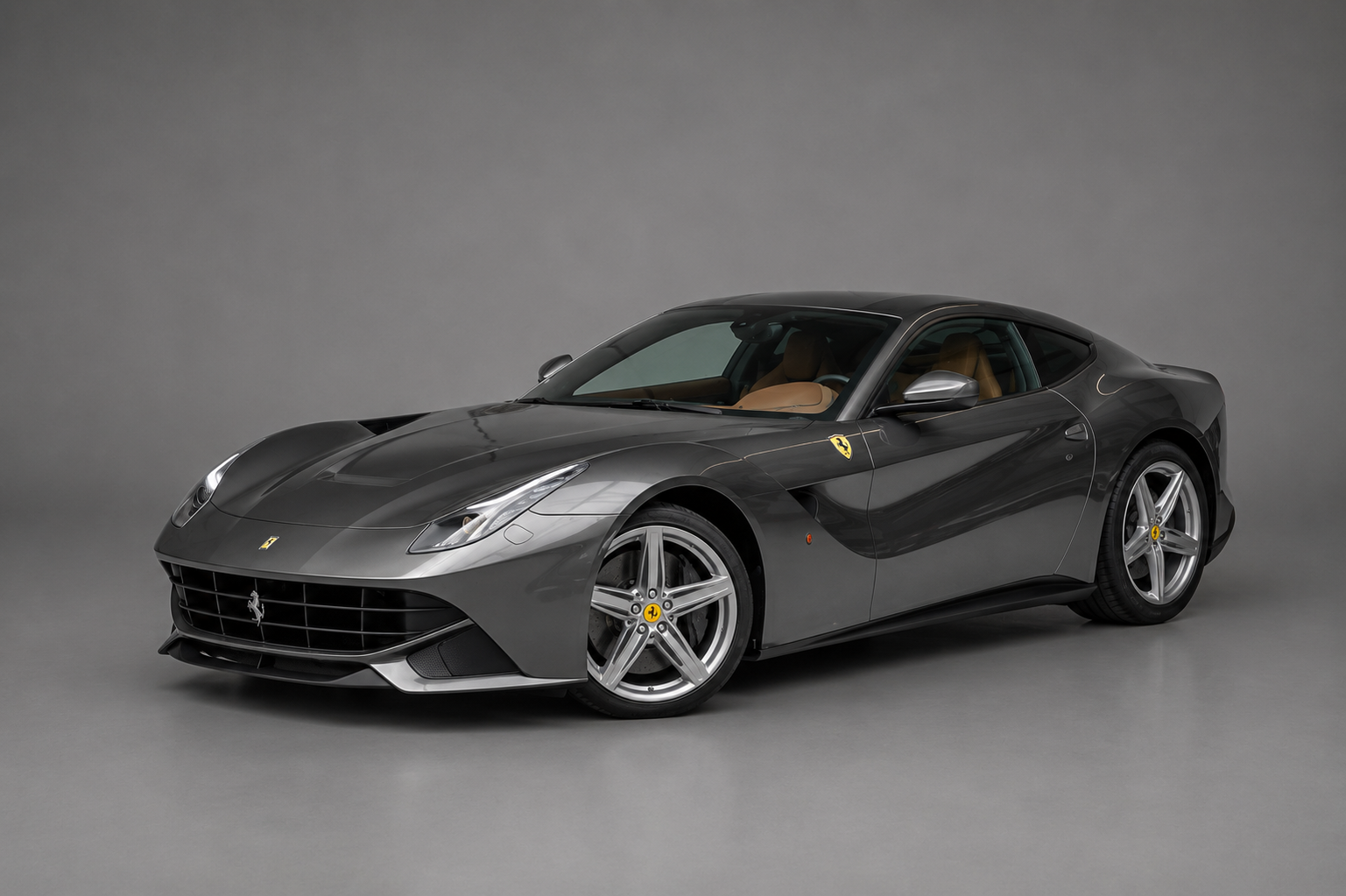 Grigio Silverstone grey Ferrari F12berlinetta coupe, front three-quarter studio view on a plain grey backdrop showing the long bonnet with aero bridge, side vents and five-spoke alloy wheels.