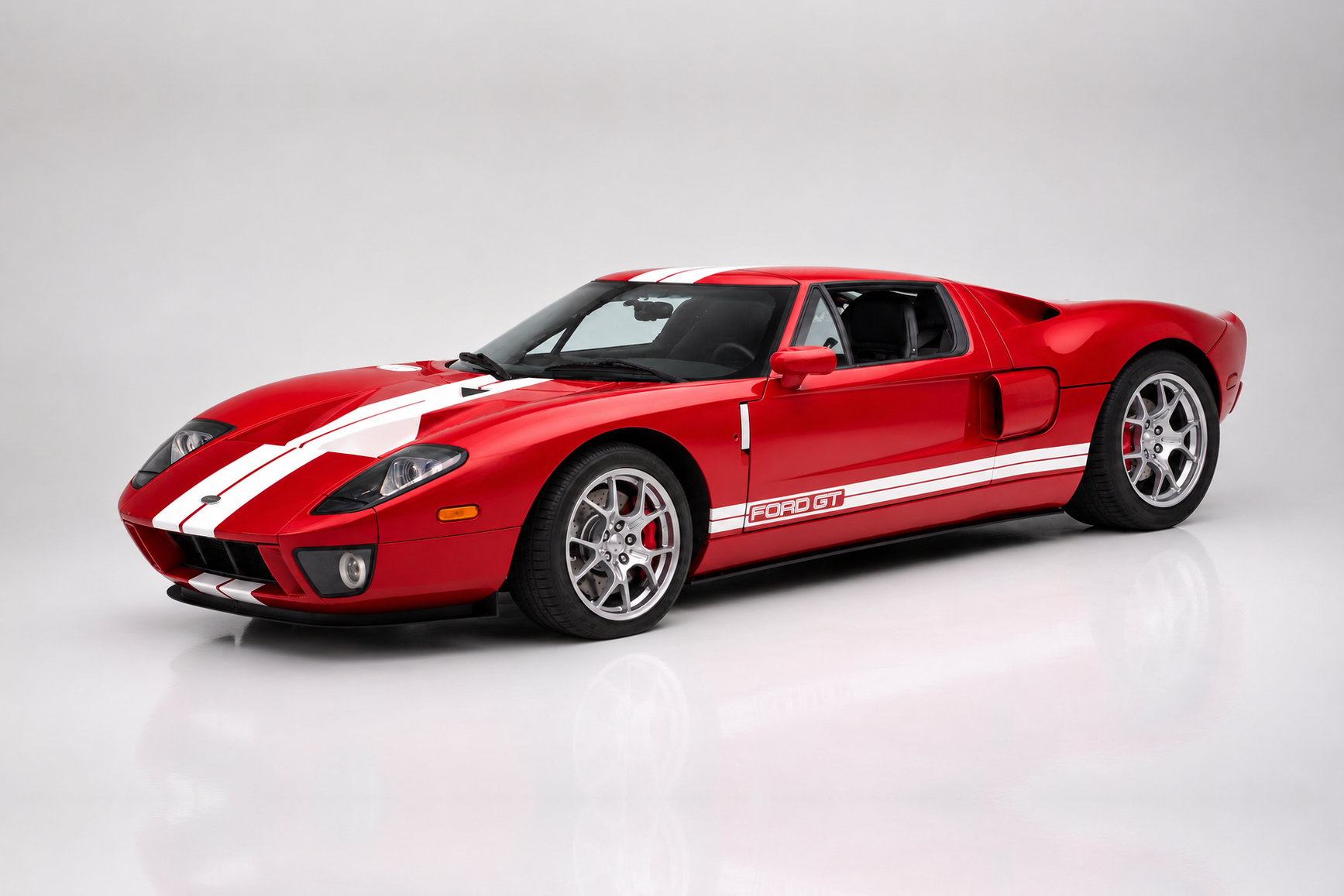 Red Ford GT (2005–2006) with twin white racing stripes in a studio setting, front three-quarter view showing the round headlights, side intake scoops, 'FORD GT' door graphic and polished five-spoke alloy wheels.