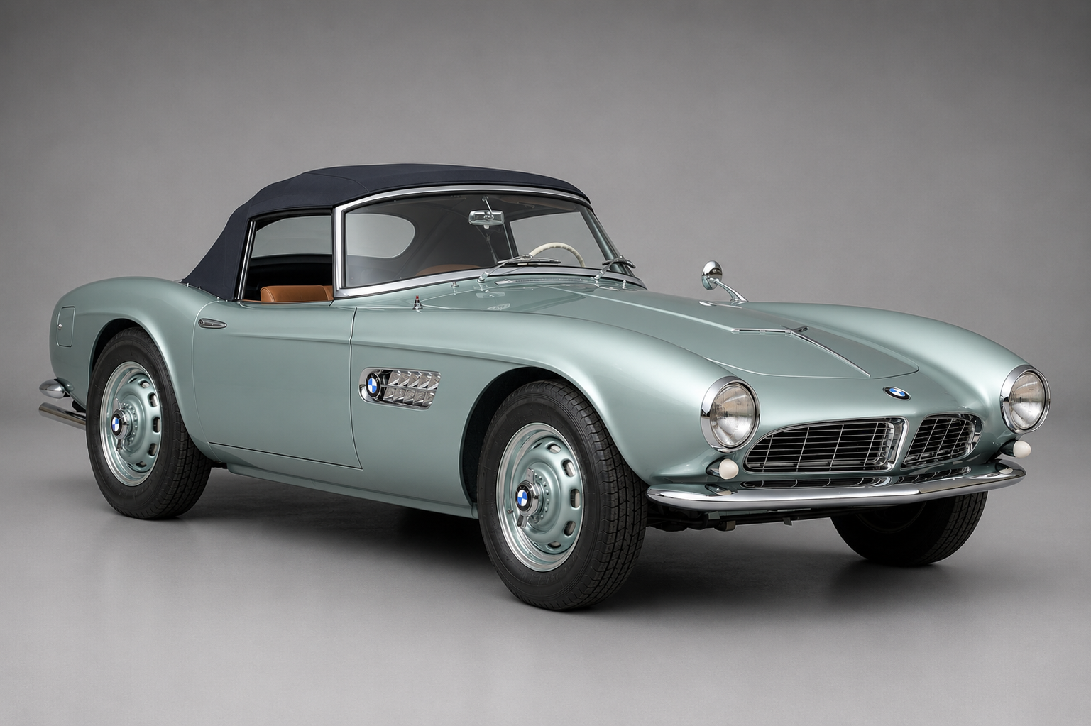 Pale silver-blue BMW 507 Roadster with a dark navy soft top raised, front three-quarter studio view on a plain grey backdrop showing the chrome bumper, rounded headlights, side vent grille and tan leather interior.