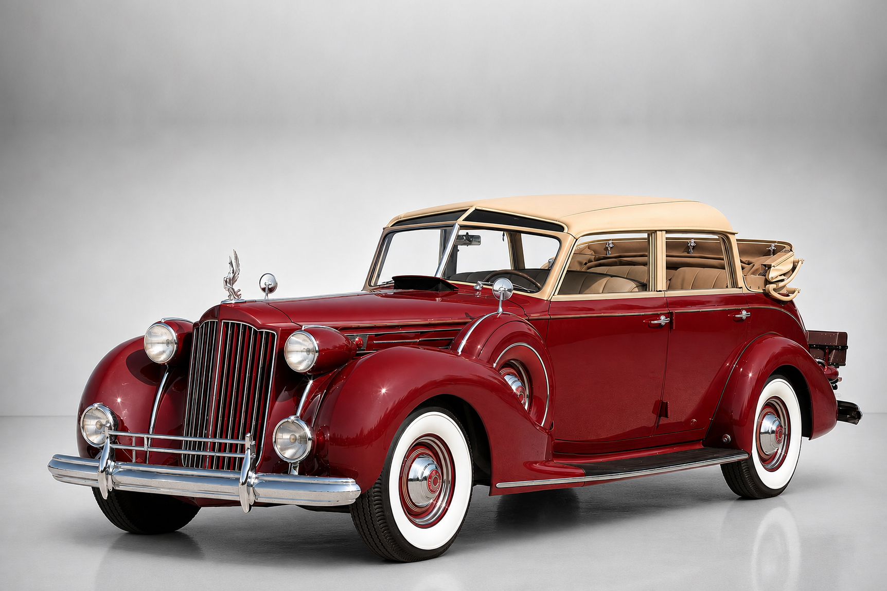 Dark red Packard Twelve convertible sedan with tan folding roof in a studio setting, front three-quarter view showing the vertical-bar chrome grille, cormorant hood ornament, dual side-mounted spare wheels, chrome bumpers with over-riders and whitewall tyres on body-colour wheels with chrome hub caps.