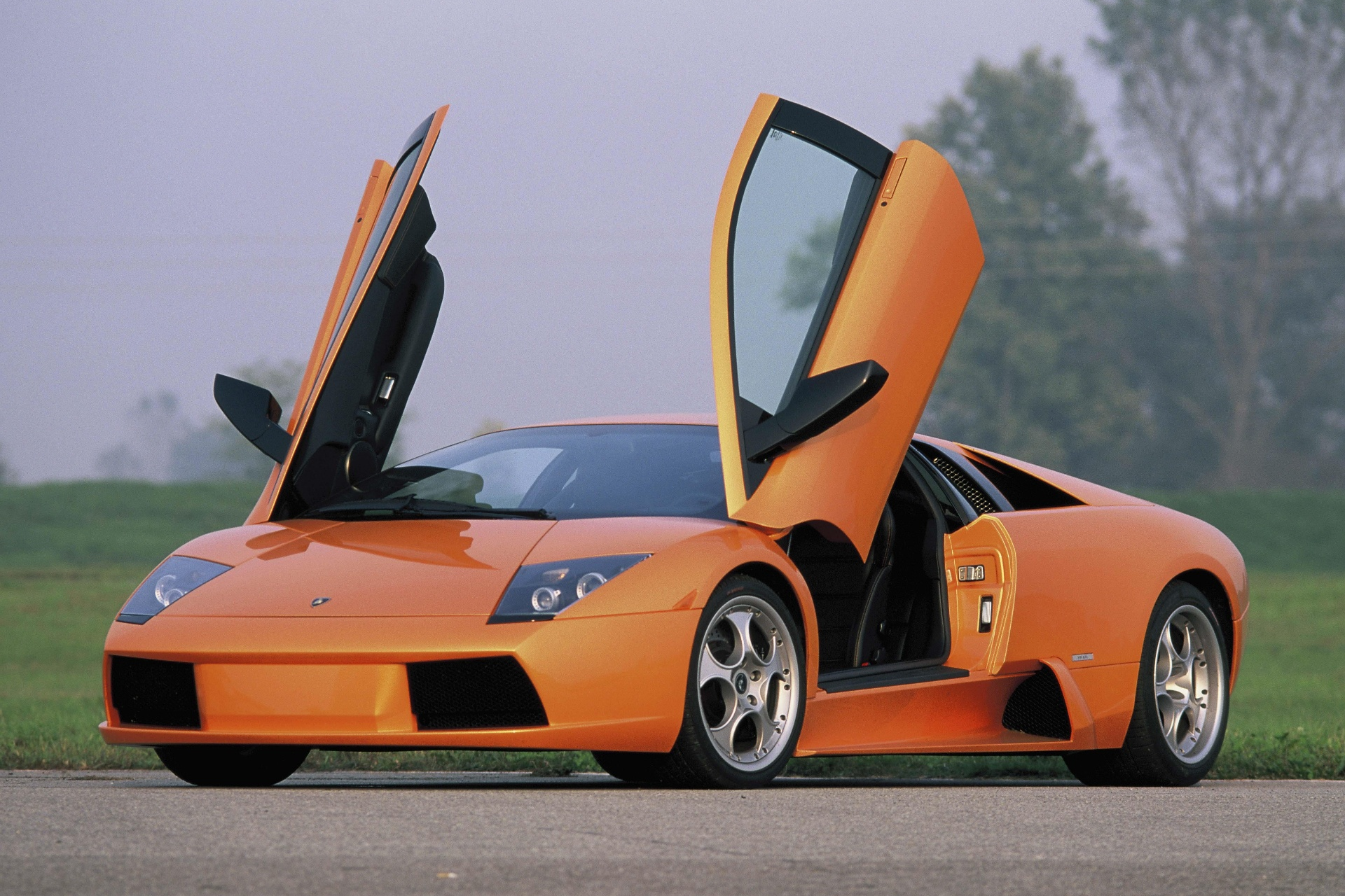 Orange Lamborghini Murciélago photographed outdoors on tarmac, front three-quarter view with both scissor doors raised, showing the first-generation bodywork, angular headlamp housings, wide low nose with black lower intakes, side air scoop and silver alloy wheels.