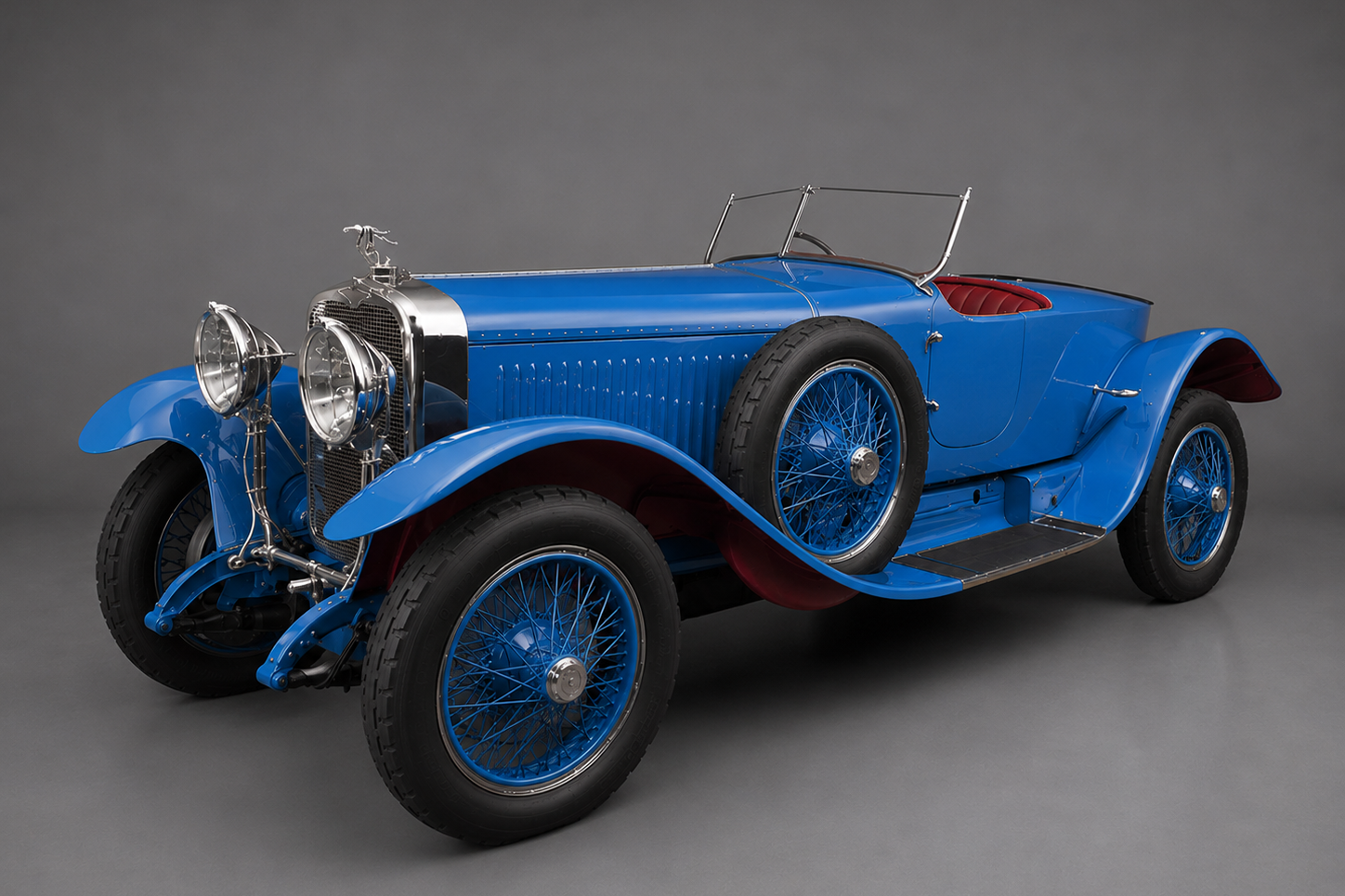 Bright blue 1920s Hispano-Suiza H6 open sports tourer with matching blue wire wheels and a red leather interior, front three-quarter studio view on a plain grey backdrop showing the polished radiator, stork mascot, twin headlamps and side-mounted spare wheel.