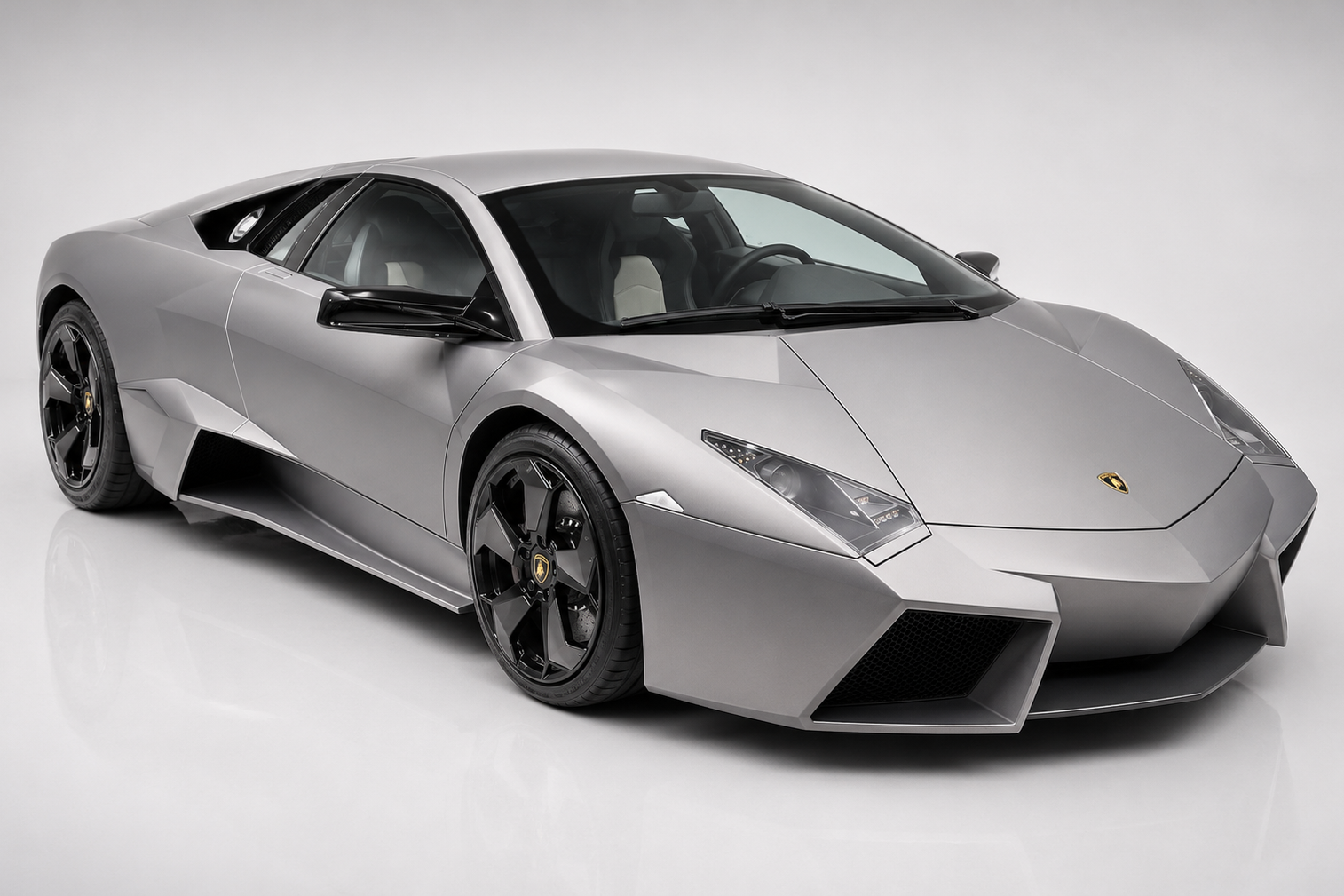 Matte grey Lamborghini Reventón coupe in a bright studio, front three-quarter view showing the stealth-fighter faceted bodywork, sharp LED headlamps, angular black lower intakes, side air scoop and black multi-spoke alloy wheels.