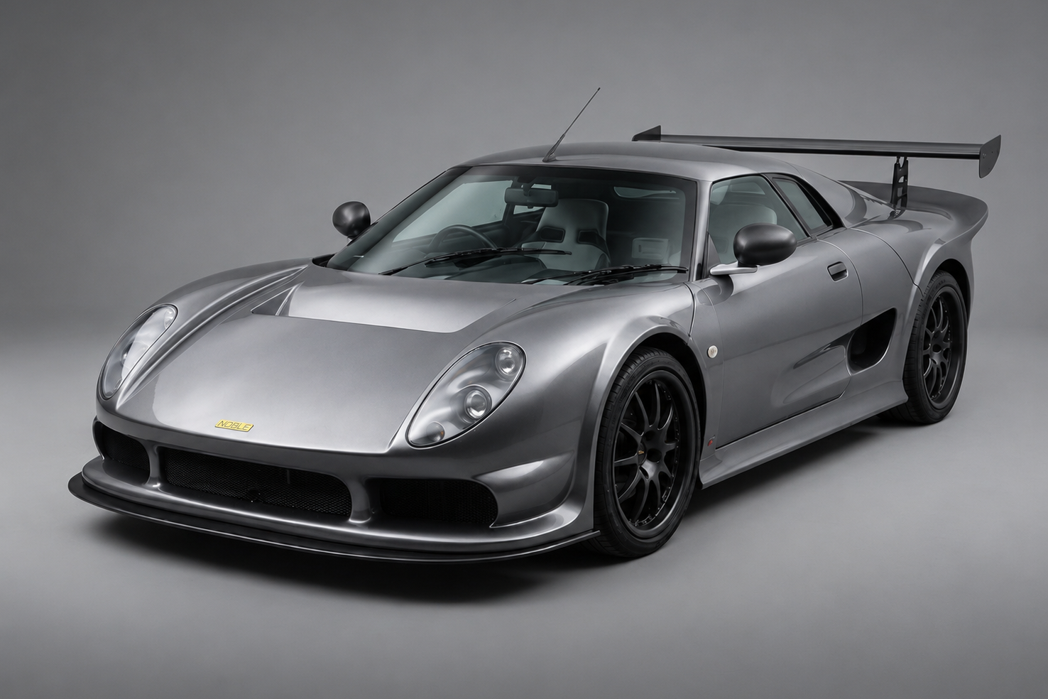 Silver-grey Noble M12 GTO-3R in a studio, front three-quarter view showing the twin headlamps under smooth covers, black pod mirrors, large rear wing, Noble badge on the nose and black multi-spoke wheels.
