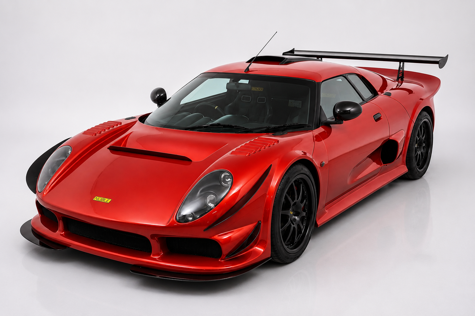 Deep metallic red Noble M400 mid-engined coupé with a large rear wing, front three-quarter view on a studio turntable, showing the covered twin headlamps, vented bonnet, side air intakes and black multi-spoke wheels.
