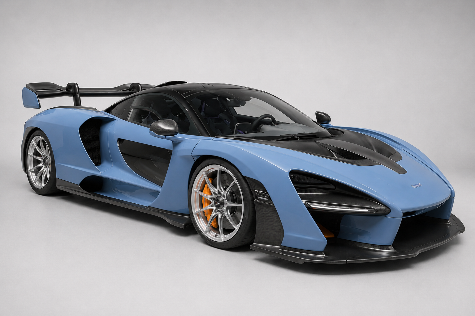Studio three-quarter front view of a blue McLaren Senna with black carbon roof, roof scoop and prominent rear wing, silver wheels and orange brake calipers on a plain grey backdrop.