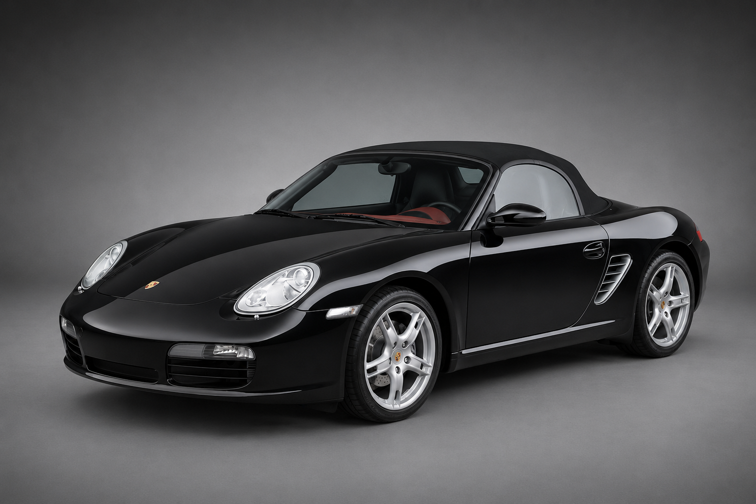 Black Porsche Boxster convertible with cloth top raised, photographed front three-quarter in a studio setting, showing silver five-spoke wheels, side air intake and glimpse of red leather interior.