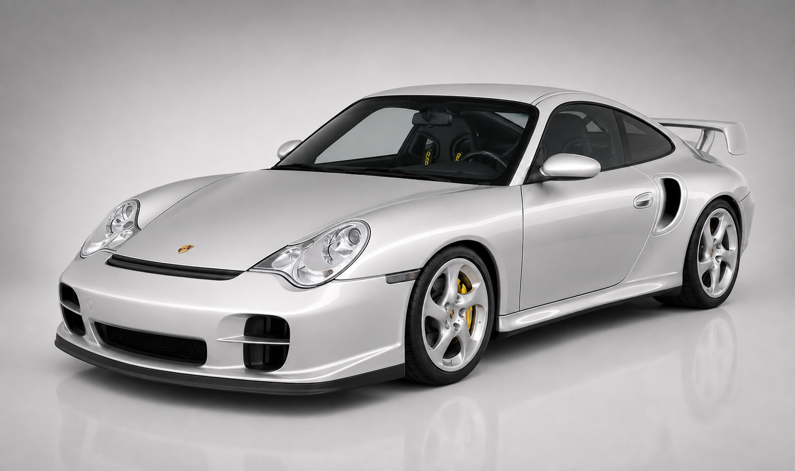 Silver Porsche 911 GT2 (996) in a studio, front three-quarter view showing the deep front bumper with brake cooling ducts, side intercooler intakes, fixed rear wing and multi-spoke silver wheels.