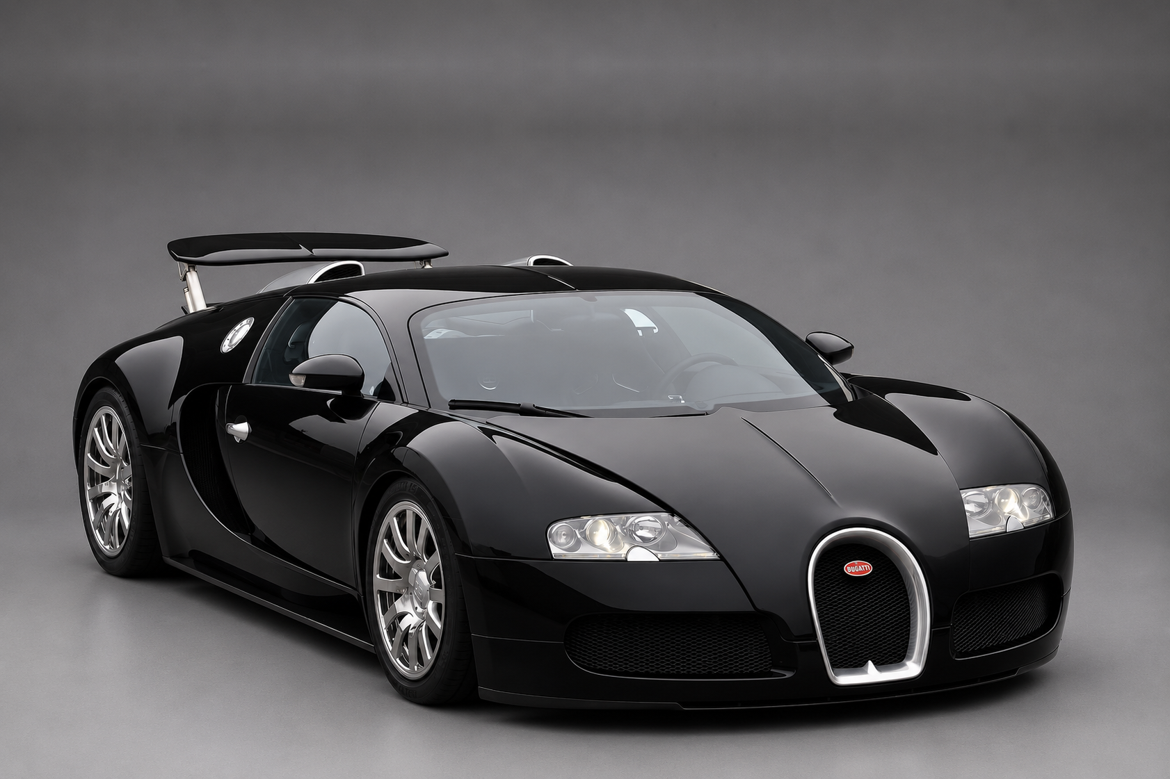 Black Bugatti Veyron 16.4 coupe with polished silver alloy wheels and a raised rear wing, front three-quarter studio view on a plain grey backdrop showing the horseshoe grille and quad round headlights.