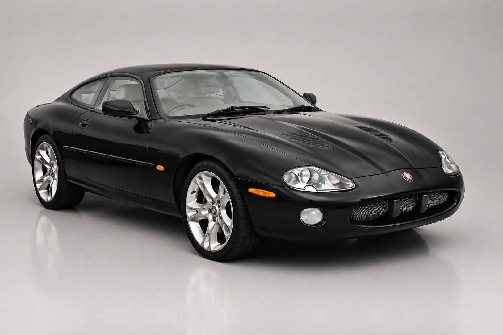 Black Jaguar XKR (X100) coupé photographed in a studio, front three-quarter view showing the twin oval headlamps, mesh oval grille with growler badge, bonnet louvres, chrome side strake and multi-spoke alloy wheels.