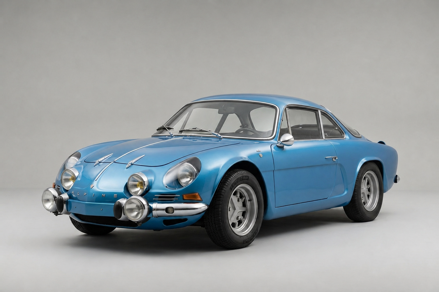 Blue Alpine A110 Berlinette in a studio, front three-quarter view showing the low fibreglass body, twin chrome-ringed headlamps with auxiliary rally spotlamps on the front panel, Alpine script on the nose and silver alloy wheels.