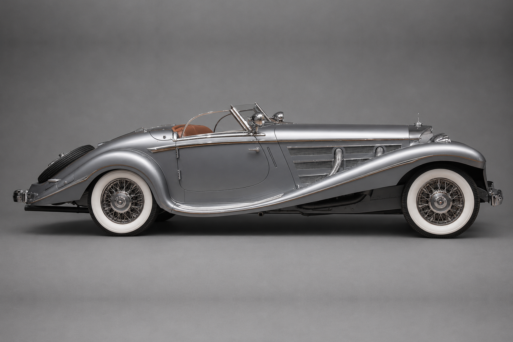 Studio side profile of a silver Mercedes-Benz 540K Special Roadster with long bonnet, sweeping front and rear fenders, whitewall tyres, wire wheels and rear-mounted spare, on a plain grey backdrop.
