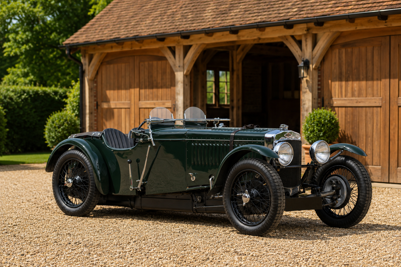 Dark green pre-war Frazer Nash TT Replica open two-seater with cycle-style front wings, aero screens, exposed side exhaust and wire wheels, parked on gravel in front of an oak-framed cart lodge.