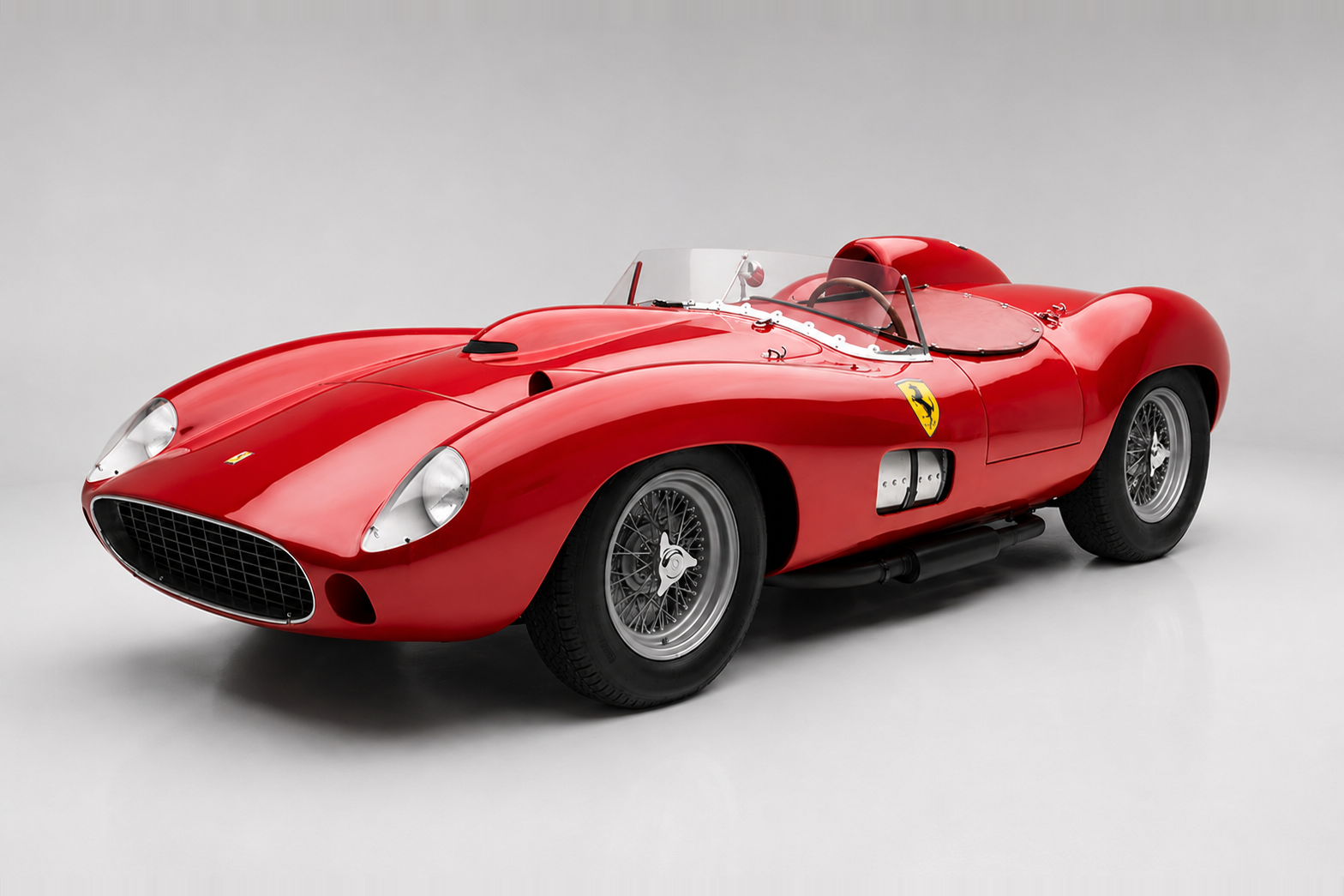 Red Ferrari 335 S barchetta photographed in a studio, front three-quarter view showing a low aluminium body by Scaglietti, twin faired-in headlamps, a small oval grille, a raised head-fairing behind the driver's seat, a low aero-screen, side-exit exhausts and Borrani-style wire wheels — a factory-yellow-shielded prancing horse on the flank.