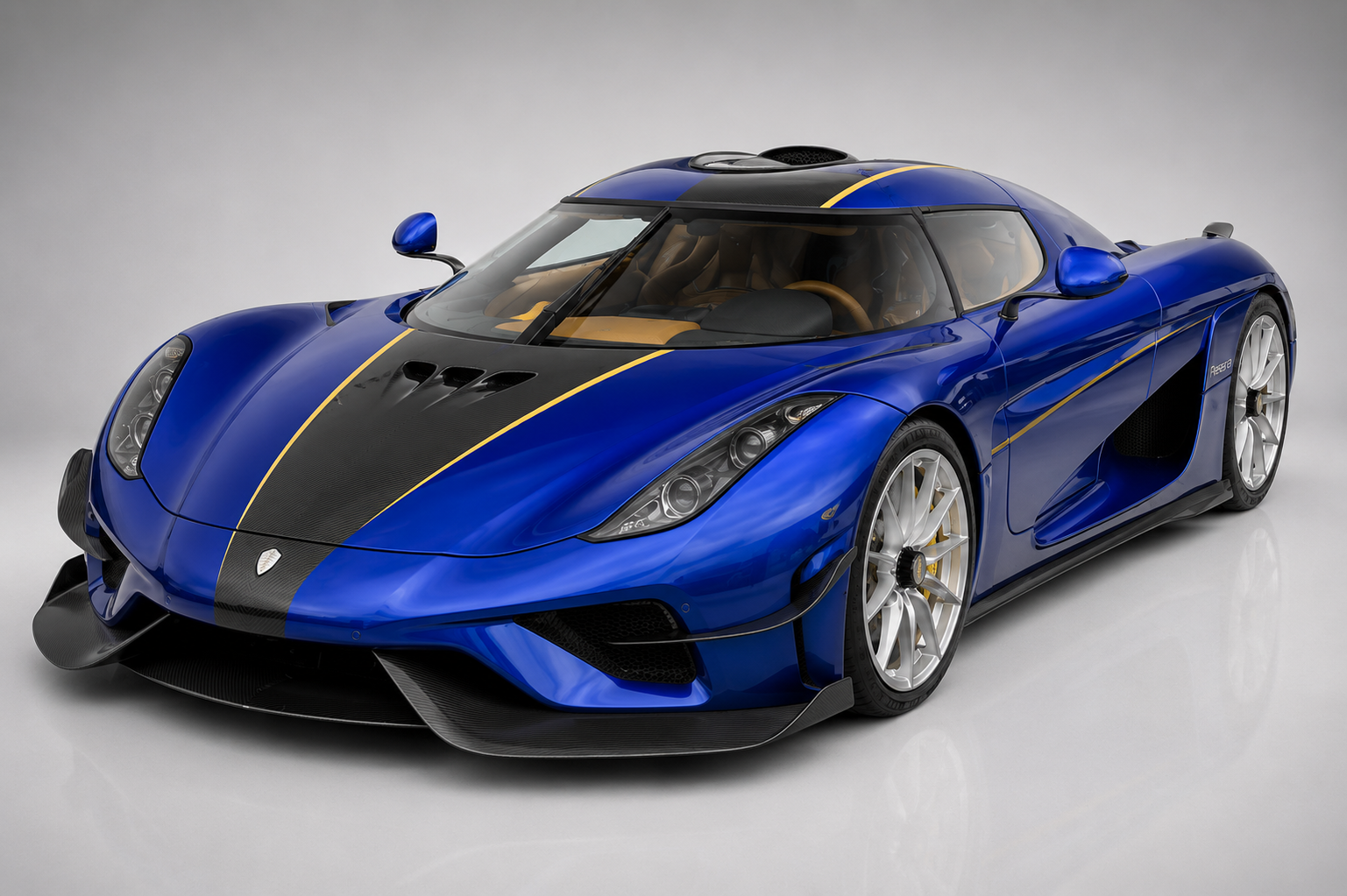 Blue Koenigsegg Regera hybrid hypercar in a studio setting, front three-quarter view showing the carbon-fibre centre stripe with gold pinstriping, distinctive Autoskin body panels, large side intakes, multi-spoke alloy wheels and the direct-drive hybrid hypercar proportions.