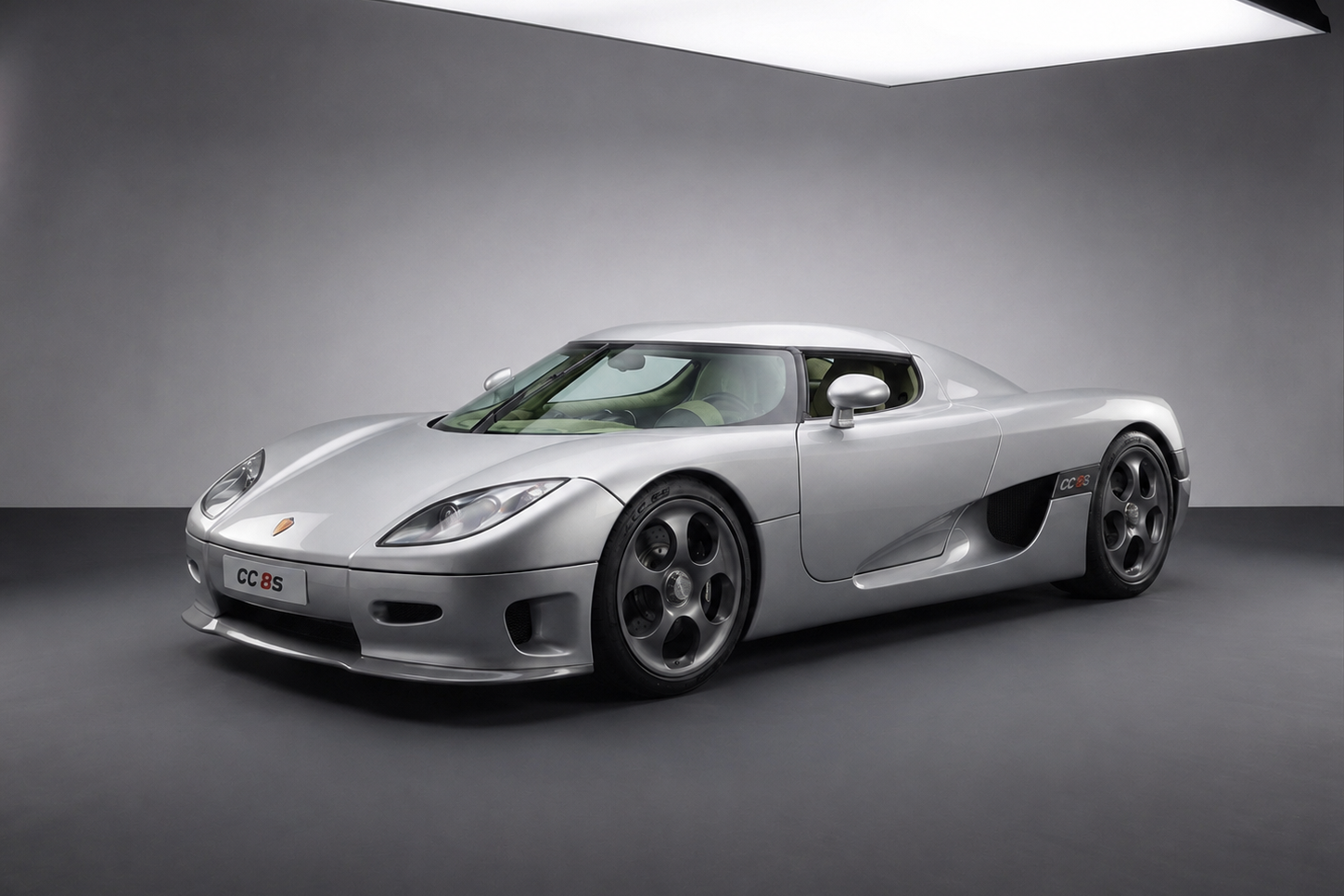 Koenigsegg CC8S in silver, front three-quarter studio view under an overhead light, showing the low targa-roof mid-engined silhouette, dihedral-hinge door, side intake feeding the supercharged V8, five-spoke turbine-style wheels and the small CC8S badge behind the front wheel arch — the founding production Koenigsegg.