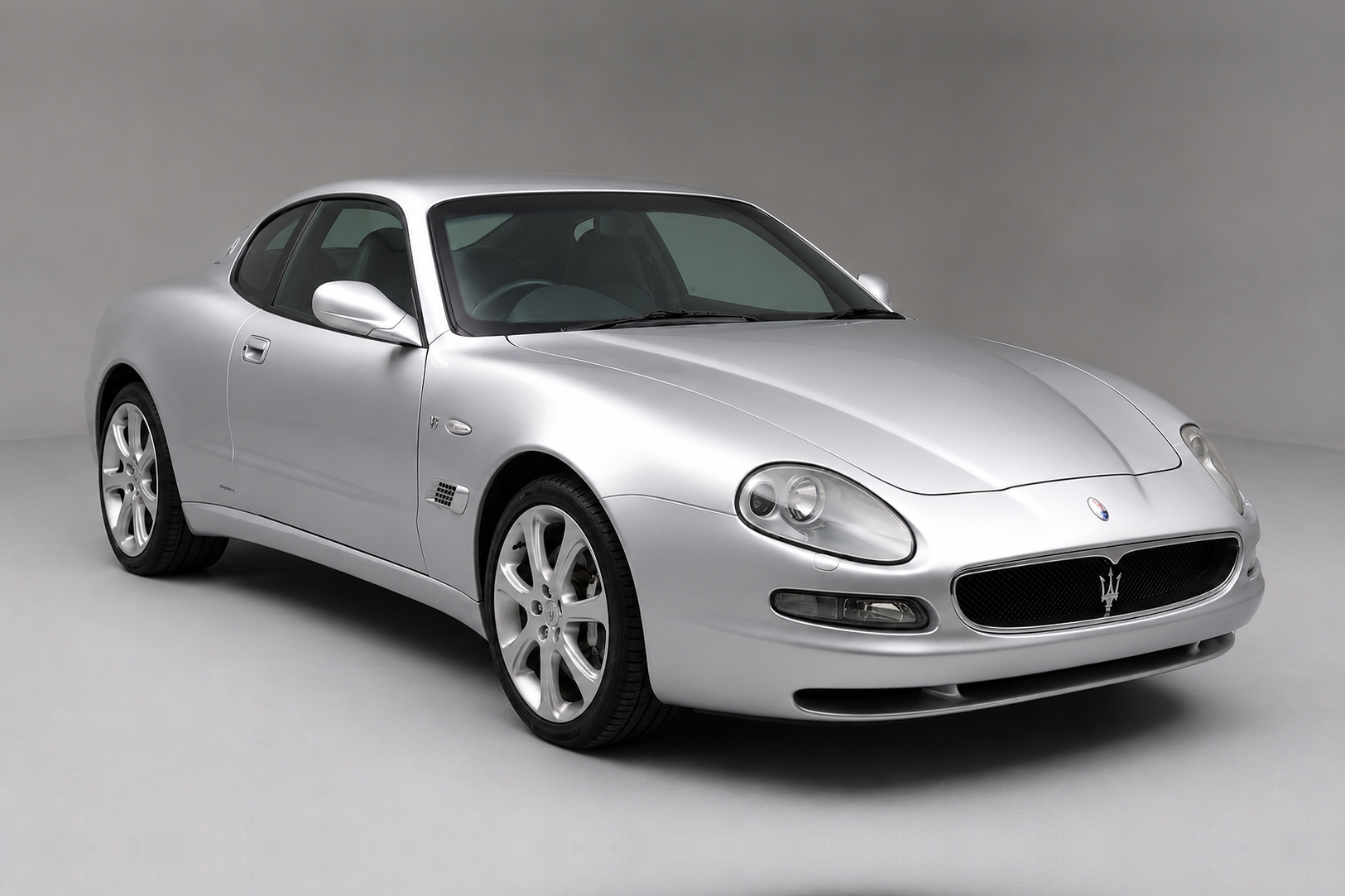 Silver Maserati 4200 Coupé photographed in a studio, front three-quarter view showing the Giugiaro body, oval Trident grille, quad headlamps, side vent and multi-spoke alloy wheels.
