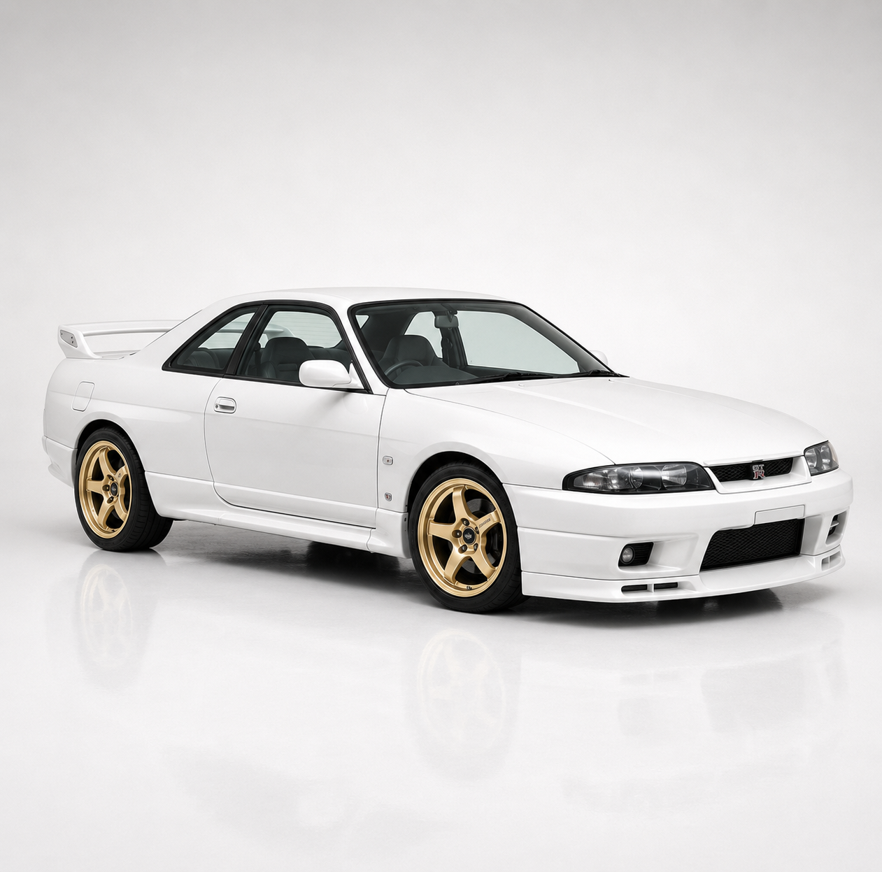 White Nissan Skyline GT-R R33 in a studio setting, front three-quarter view showing the smoother, larger 1990s coupe body, twin round headlamps, tall rear wing and gold multi-spoke wheels.