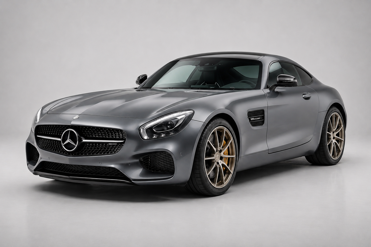 Grey Mercedes-AMG GT coupe in a plain studio setting, front three-quarter view showing the diamond-mesh grille with central three-pointed star, LED headlights, side vent badging and bronze multi-spoke alloy wheels with yellow brake calipers.