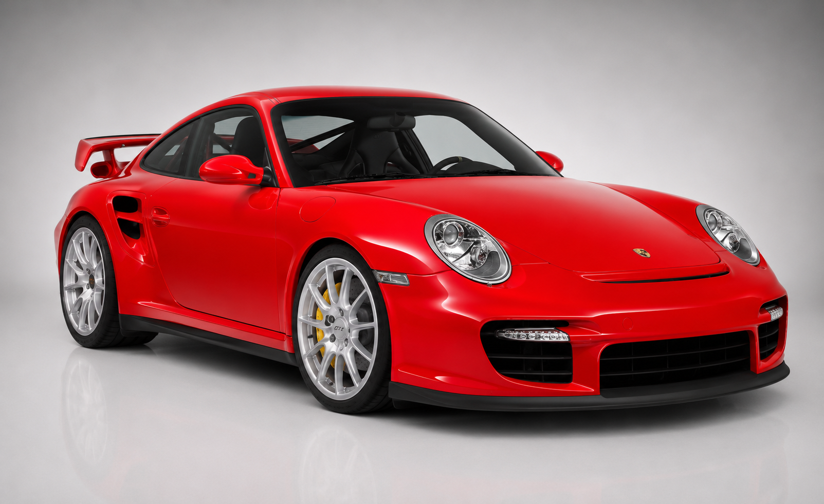 Red Porsche 911 GT2 (997) in a studio, front three-quarter view showing the deep front splitter with LED daylight running lights, side intercooler intakes, fixed rear wing and forged silver wheels.