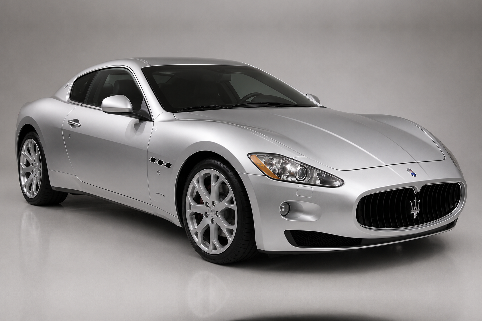Silver Maserati GranTurismo coupé photographed in a studio, front three-quarter view showing the Pininfarina 2+2 silhouette, concave oval Trident grille, quad headlamps, side vents and multi-spoke alloy wheels.