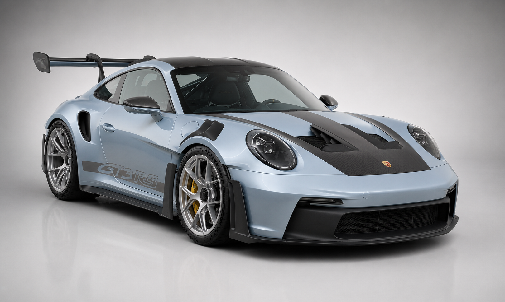 Light-blue Porsche 911 GT3 RS (992) in a grey studio, front three-quarter view showing the DRS-equipped swan-neck rear wing, exposed carbon bonnet with central duct and silver forged wheels.