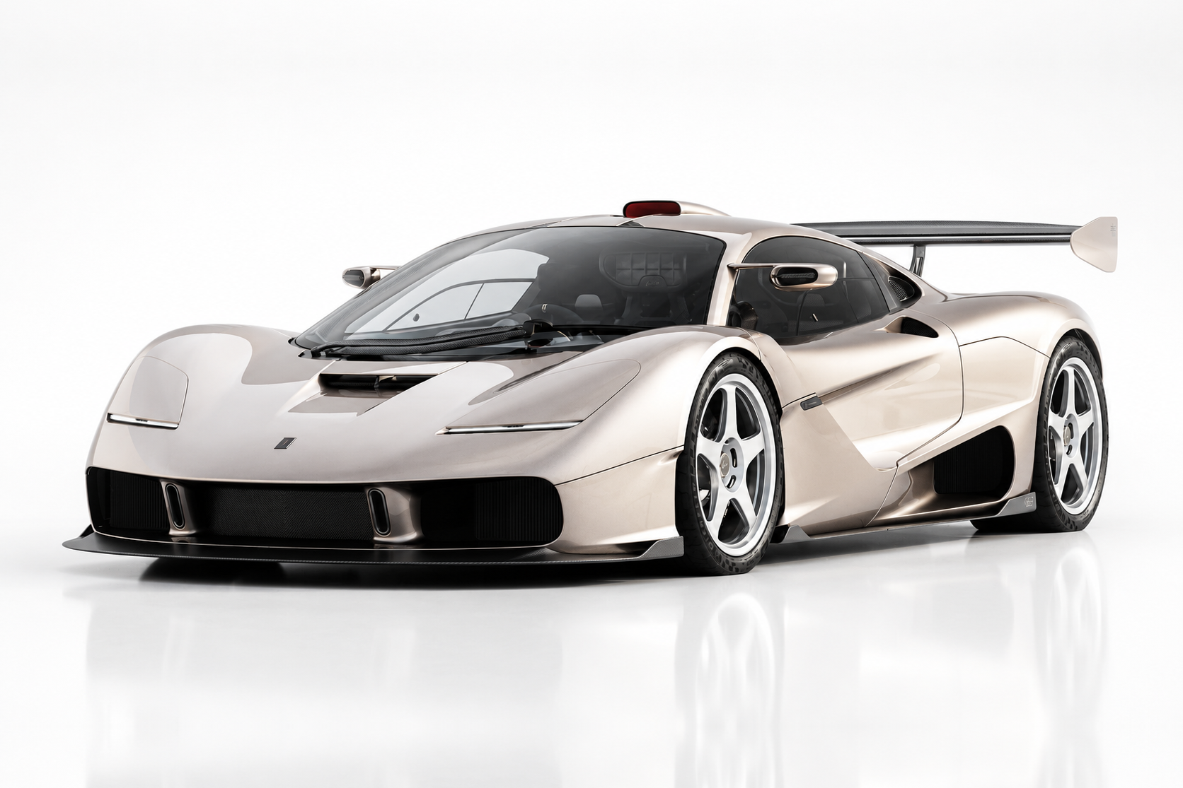 Champagne-silver Gordon Murray Automotive S1 LM photographed in a studio, front three-quarter view showing the McLaren F1-inspired silhouette, low nose, roof-mounted intake snorkel, tall rear wing, exposed carbon underbody and five-spoke wheels.