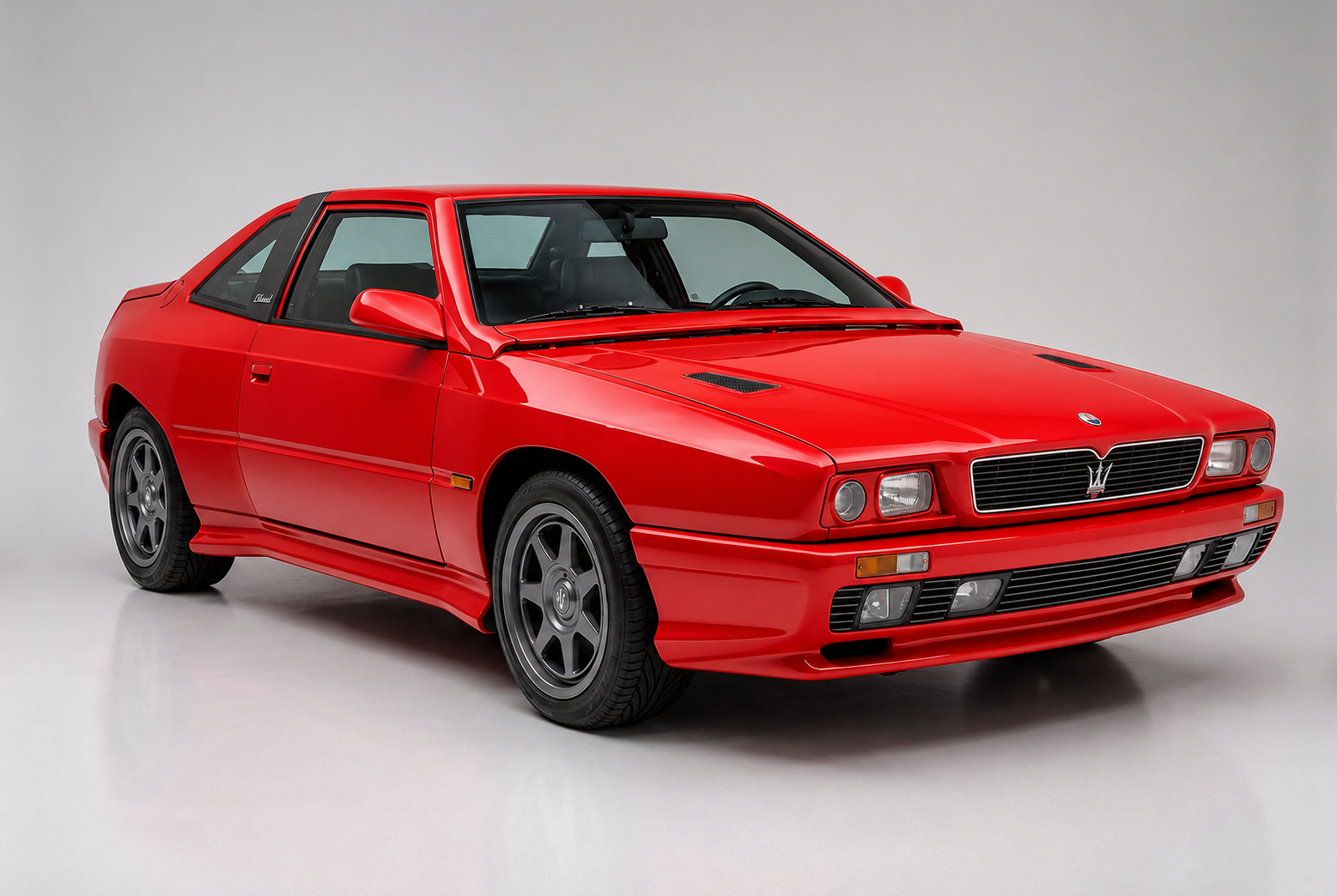 Red Maserati Shamal coupé photographed in a studio, front three-quarter view against a neutral grey background, showing the Gandini-designed Biturbo-era nose with twin round headlamps, the Maserati trident grille, side vent and multi-spoke alloy wheels.