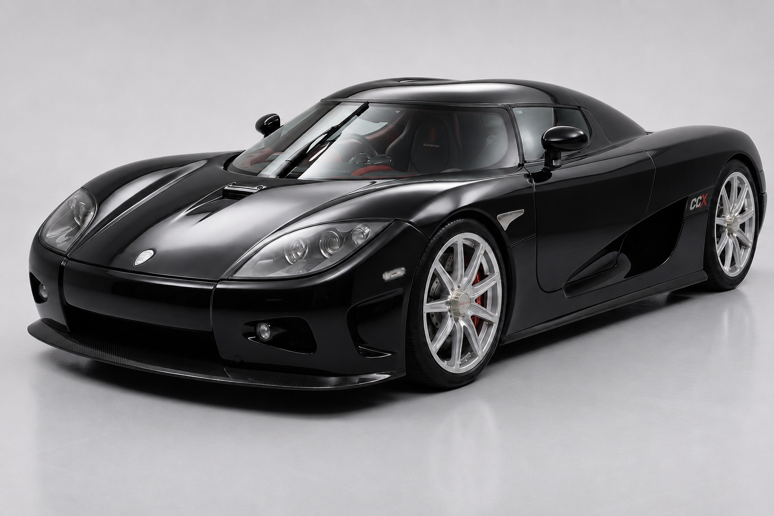 Koenigsegg CCX in gloss black with exposed carbon-fibre front splitter, red 'CCX' side badge and polished multi-spoke wheels with red brake calipers, front three-quarter studio view showing the low mid-engined silhouette, dihedral synchro-helix door and side intake.