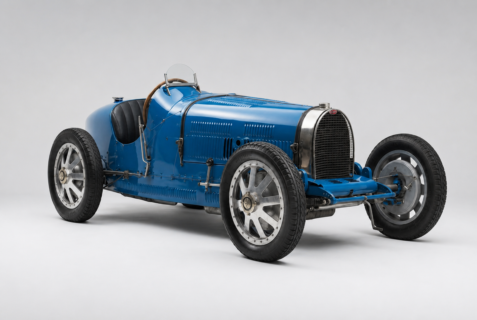 French racing blue Bugatti Type 35 Grand Prix single-seater on a plain light-grey studio backdrop, front three-quarter view showing the polished horseshoe radiator with mesh grille, aero screen, black leather seat, wooden steering wheel, exposed front axle and suspension, and cast alloy wheels.
