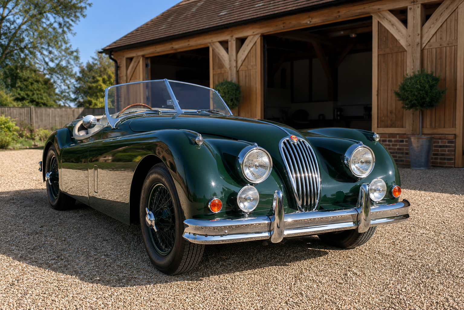 British racing green Jaguar XK roadster with the top down and chrome wire wheels, front three-quarter view parked on gravel in front of a timber-framed barn on a sunny day.