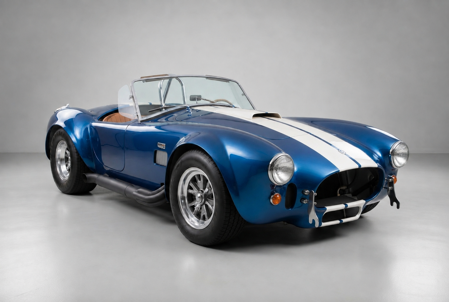 Blue AC Cobra roadster with a white centre racing stripe, front three-quarter studio view on a plain grey backdrop showing the open cockpit with twin aero screens, polished five-spoke alloy wheels, side-exit exhaust and round headlights.
