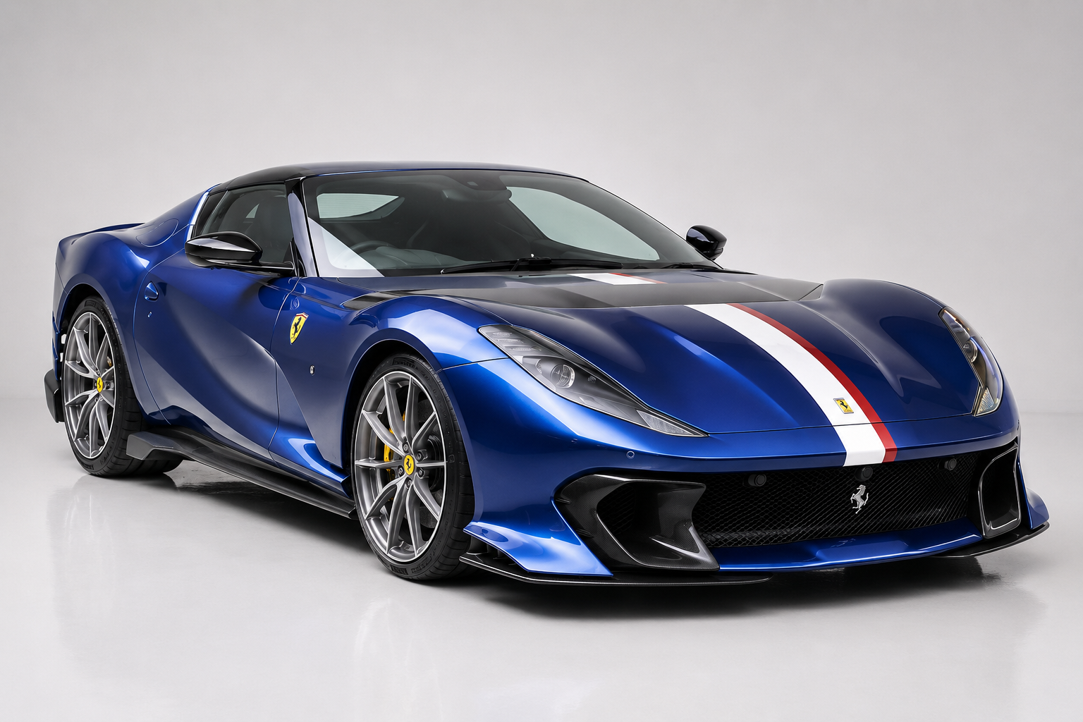 Blue Ferrari 812 Competizione in a studio setting, front three-quarter view showing the Italian tricolore stripe over the bonnet and roof, exposed carbon roof and front splitter, yellow brake calipers and diamond-cut alloy wheels.