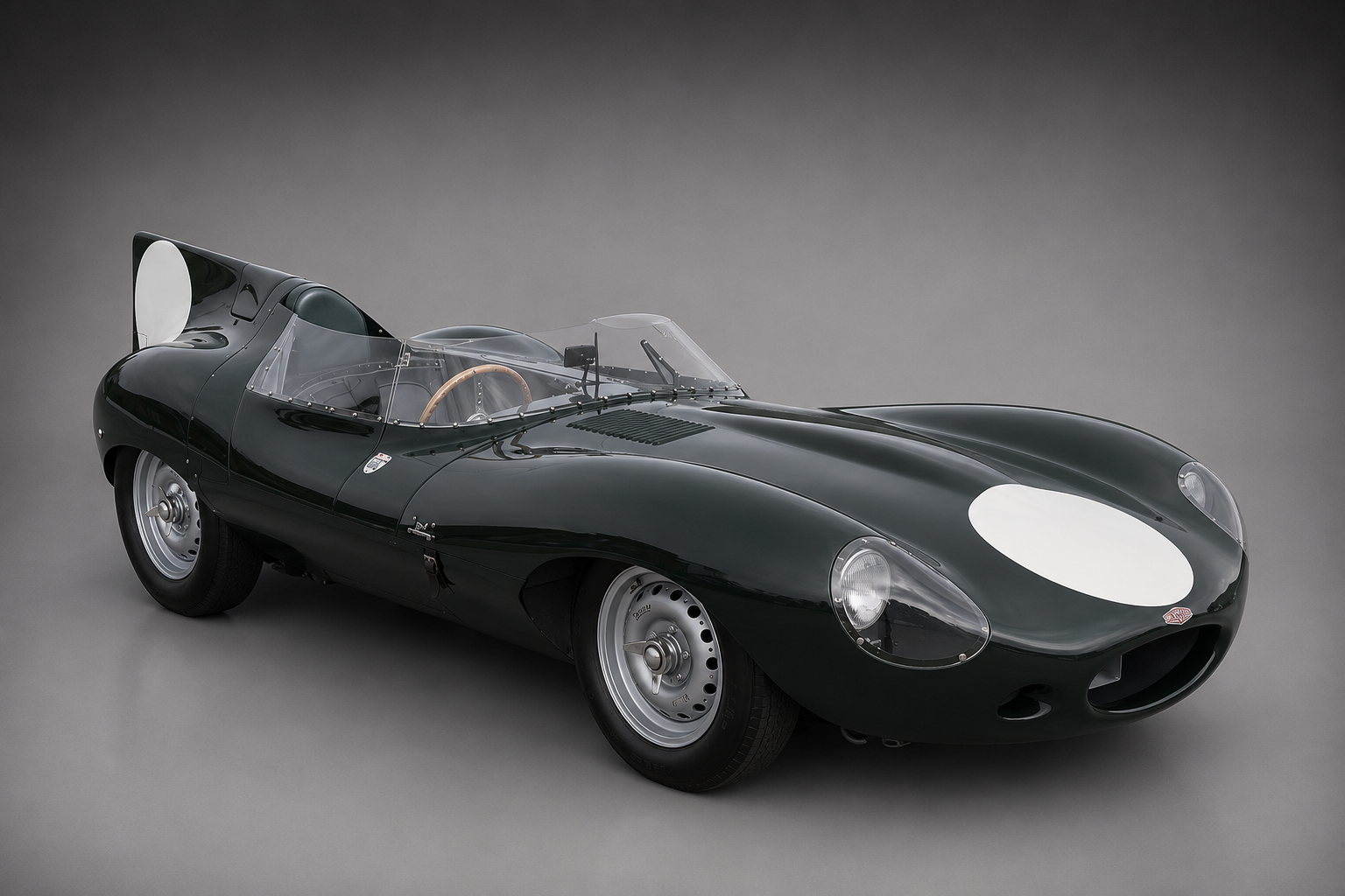 British racing green Jaguar D-Type racing car with tall rear fin and white roundels on the nose and flank, front three-quarter studio view on a plain grey backdrop showing the low aerodynamic bodywork and open cockpit.