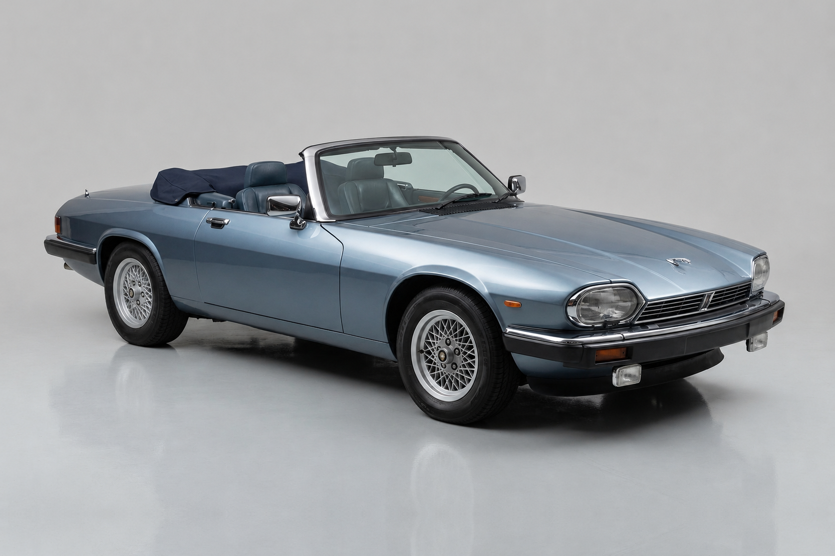 Light blue Jaguar XJS convertible photographed in a studio, front three-quarter view with the roof down and tonneau cover fitted, showing the flush headlamps, chrome grille surround, side crease, mesh-style alloy wheels and long-bonnet grand-tourer proportions.