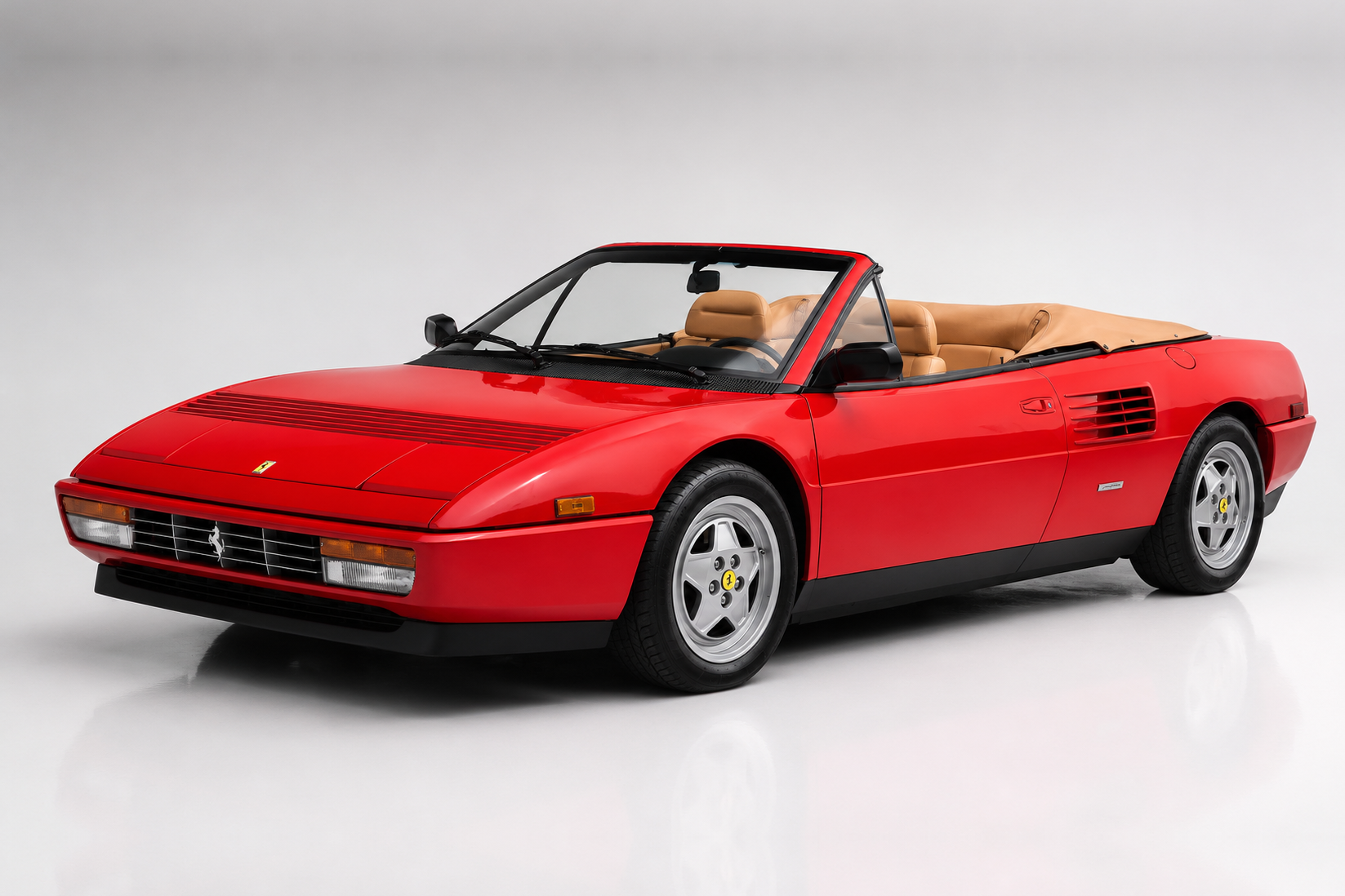 Red Ferrari Mondial Cabriolet with tan leather interior and folded soft-top, front three-quarter studio view against a light grey background showing the flush rectangular headlamps, black grille with prancing horse badge, side air intake ahead of the rear wheel and silver five-spoke alloy wheels with yellow prancing-horse centres.