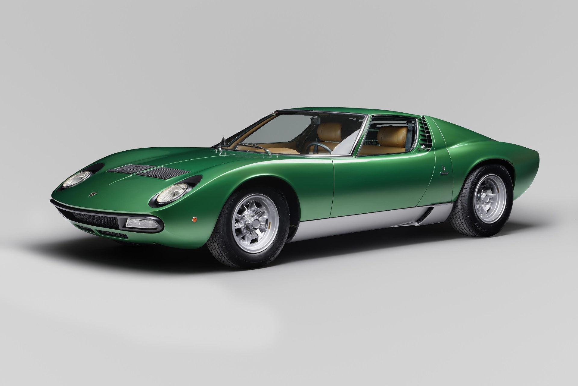 Green Lamborghini Miura P400 SV in a bright studio, front three-quarter view showing the low mid-engined Bertone berlinetta silhouette, headlamps without the earlier 'eyelash' surrounds, widened rear arches, side air intakes and polished five-spoke Campagnolo alloy wheels.