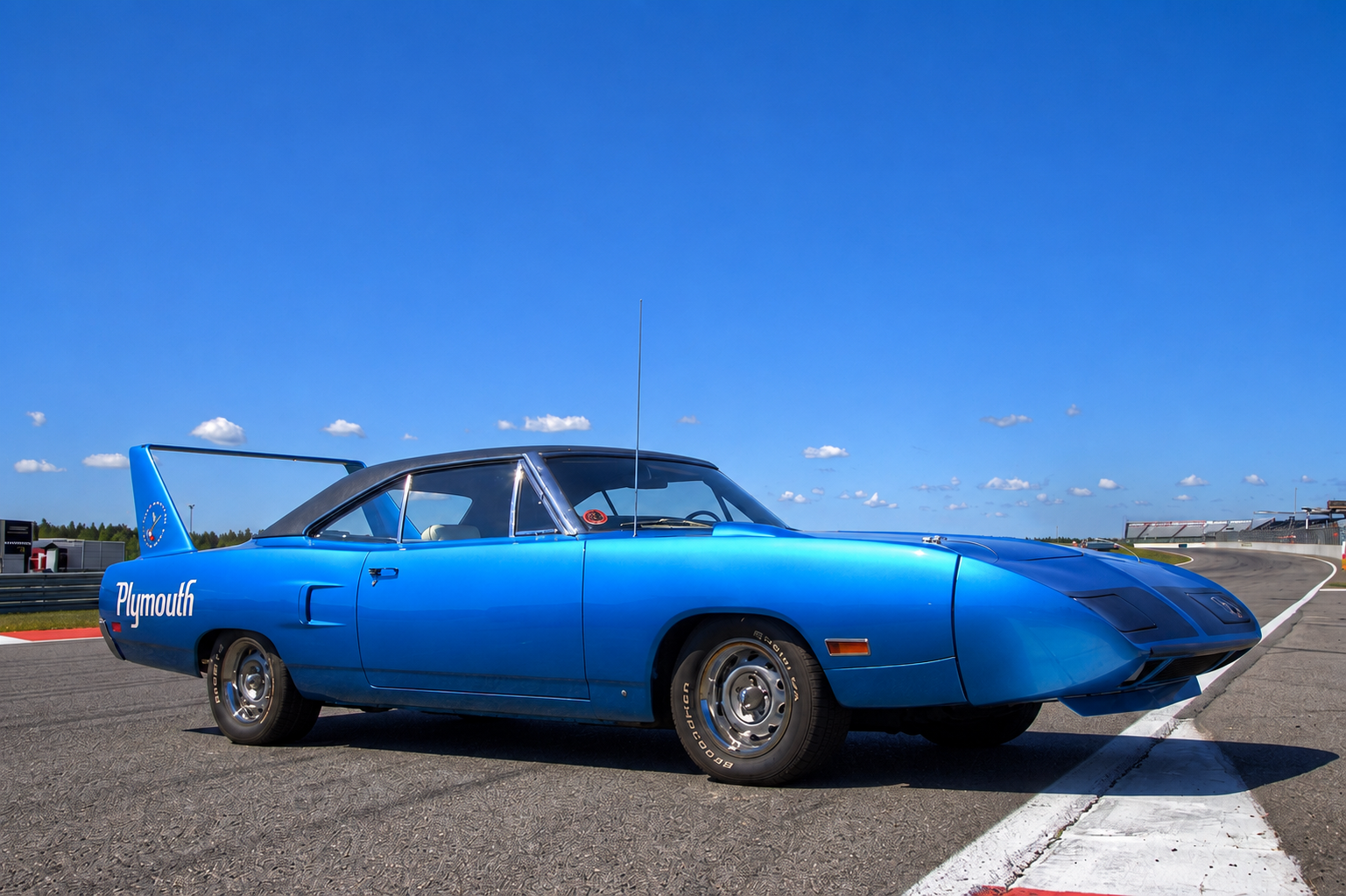 Plymouth Superbird in Petty Blue with black vinyl roof on a race circuit, rear three-quarter view showing the fully-extended fibreglass nose cone at the far side, the tall aluminium-uprighted rear stabiliser wing rising well above the roofline, 'Plymouth' script on the rear quarter panel and steel wheels with trim rings — Chrysler's 1970 NASCAR aero homologation built on the Road Runner platform.