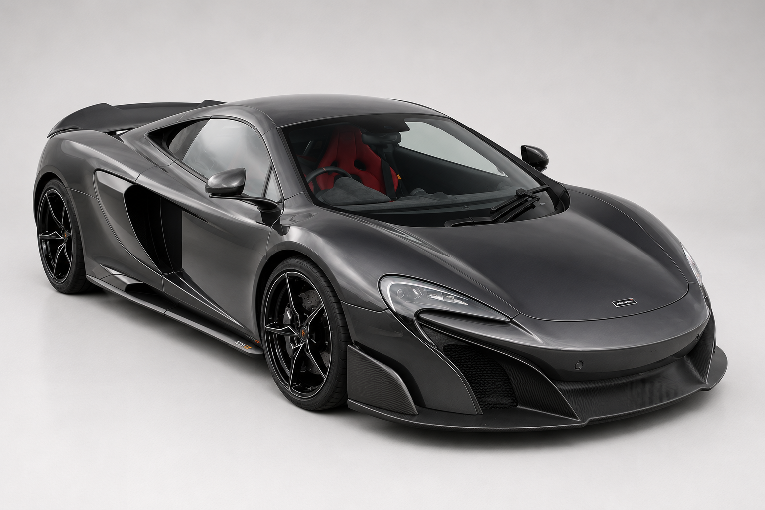Dark grey McLaren 675LT in a studio setting, front three-quarter view showing its extended carbon aero, dark alloy wheels and red interior accents.