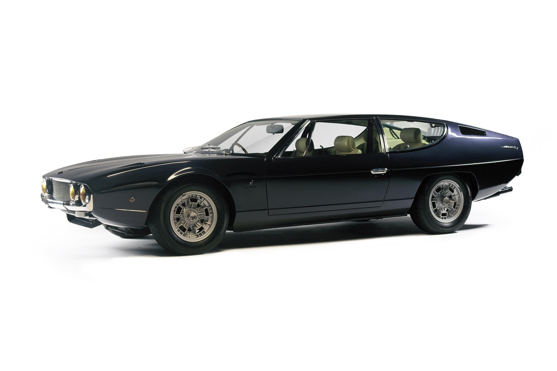 Dark blue Lamborghini Espada in a bright studio, front three-quarter view showing Gandini's long low four-seat Bertone body, quad round headlamps behind a mesh grille, slim chrome bumper, sharp shoulder line and grey five-spoke alloy wheels.
